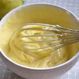 Una ciotola bianca piena di soffice crema al limone, con una frusta che la sta mescolando. Sullo sfondo, un limone intero.
