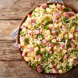 Un piatto di pasta con prosciutto cotto, piselli e formaggio grattugiato su un tavolo di legno rustico.