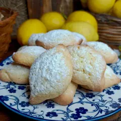 Biscotti con farina di mandorle spolverati di zucchero a velo, su un piatto decorato con fiori blu. Sullo sfondo limoni e un cestino.