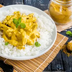 Piatto di riso bianco con straccetti di pollo al curry cremoso, guarnito con menta fresca. Un cucchiaio di curry in polvere e un vasetto completano la scena.
