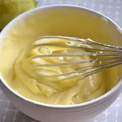 Una ciotola bianca piena di soffice crema al limone, con una frusta che la sta mescolando. Sullo sfondo, un limone intero.