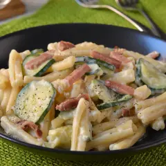 Piatto di pasta cremosa con zucchine a rondelle e cubetti di speck croccante, guarnito con pepe nero macinato.