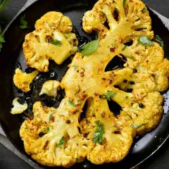 Fette di cavolo al forno dorate e speziate, servite su una padella nera con erbe fresche e pepe nero.