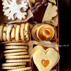 Scatola piena di biscotti classici: a fiocco di neve, con gocce di cioccolato, ripieni di caramello, a forma di cuore e ricoperti di cioccolato.