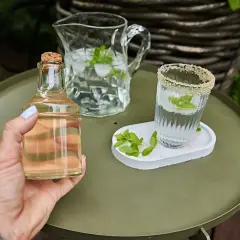 Mano tiene una bottiglietta di sciroppo di menta, accanto a un bicchiere e una caraffa con acqua, ghiaccio e foglie di menta.