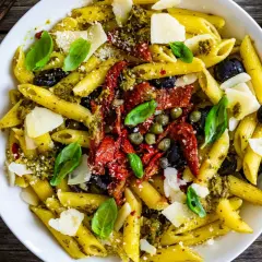 Penne al pesto con pomodori secchi, olive nere, capperi e scaglie di parmigiano. Un piatto colorato e saporito.