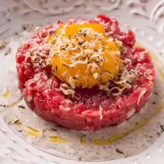 Deliziosa tartare di manzo finemente tritata, guarnita con tuorli d'uovo dorati e scaglie di formaggio, servita su un piatto elegante con una salsa cremosa.