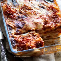 Lasagne alla bolognese appena sfornate, dorate e filanti, in una teglia di vetro. Accanto, due coltelli d'argento.