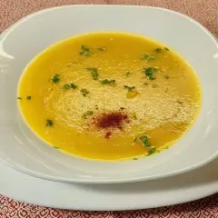 Una vellutata crema di peperoni, guarnita con prezzemolo fresco e una spolverata di paprika, servita in una ciotola bianca su un tovagliolo rosso.