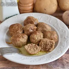 Polpette in umido succulente su un piatto bianco, con una forchetta pronta ad assaggiarle. Sullo sfondo, patate e aglio.