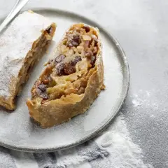 Due fette di strudel di mele con pasta sfoglia, cosparse di zucchero a velo, su un piatto grigio con una forchetta.