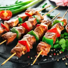 Spiedini di carne succulenti alternati a peperoni rossi e verdi, cipolla bianca, serviti su una pietra scura con pomodorini e salsa.