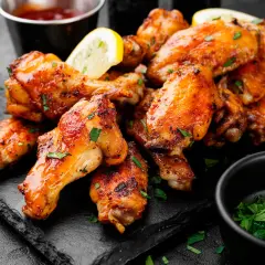 Alette di pollo dorate e succulente, guarnite con prezzemolo fresco e spicchi di limone, servite su una pietra scura con salse a lato.