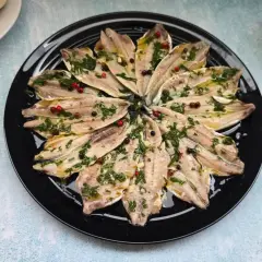Alici marinate ricetta: filetti di acciughe conditi con erbe aromatiche, aglio e pepe in grani su un piatto nero.