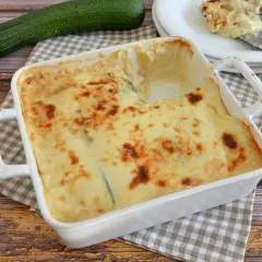 Parmigiana di zucchine bianca senza besciamella, dorata e filante, pronta per essere gustata. Accanto, una zucchina fresca.