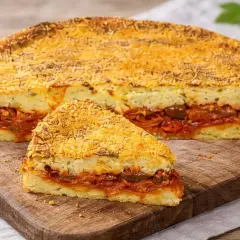 Una fetta di pitta di patate, con strati di purè e un ricco ripieno di pomodoro e olive, su un tagliere di legno.