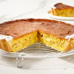 Torta di mele con crema pasticcera, con una fetta tagliata, cosparsa di zucchero a velo su una griglia metallica.