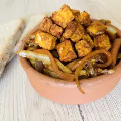 Tofu in padella dorato e saporito, servito con cipolle caramellate in una ciotola di terracotta, accanto a fette di pane rustico.