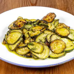 Zucchine alla poverella, fette sottili e dorate, condite con olio e erbe aromatiche, su un piatto bianco con bordo blu.