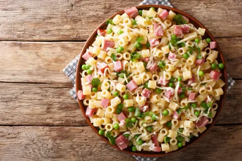 Un piatto di pasta con prosciutto cotto, piselli e formaggio grattugiato su un tavolo di legno rustico.