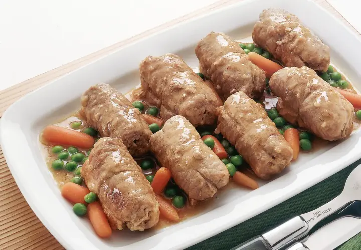 Sette involtini di carne succulenti in un piatto bianco con piselli e carote.