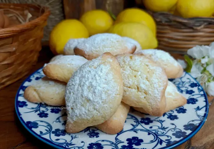 Biscotti con farina di mandorle spolverati di zucchero a velo, su un piatto decorato con fiori blu. Sullo sfondo limoni e un cestino.