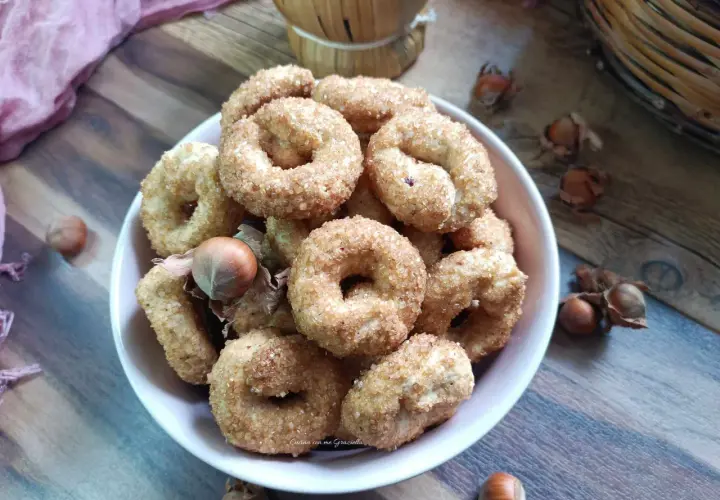 Ciambelline al vino cosparse di zucchero, con nocciole e un drappo rosa su un tavolo di legno.