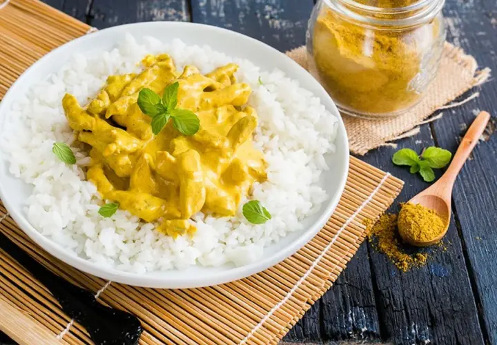 Piatto di riso bianco con straccetti di pollo al curry cremoso, guarnito con menta fresca. Un cucchiaio di curry in polvere e un vasetto completano la scena.