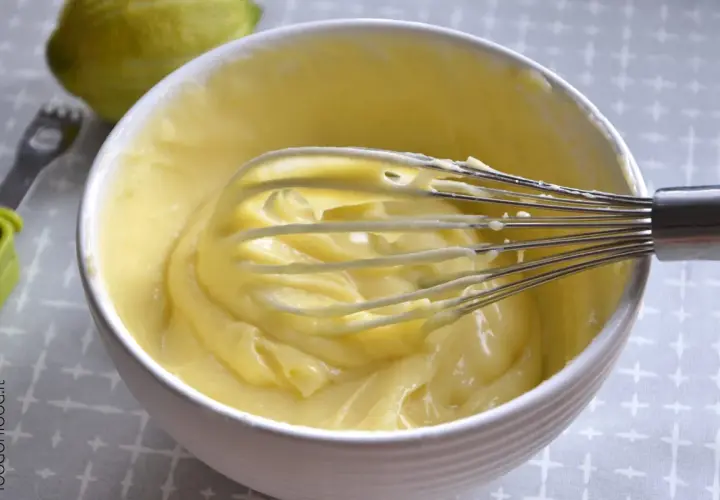 Una ciotola bianca piena di soffice crema al limone, con una frusta che la sta mescolando. Sullo sfondo, un limone intero.