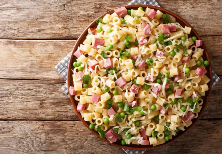 Un piatto di pasta con prosciutto cotto, piselli e formaggio grattugiato su un tavolo di legno rustico.