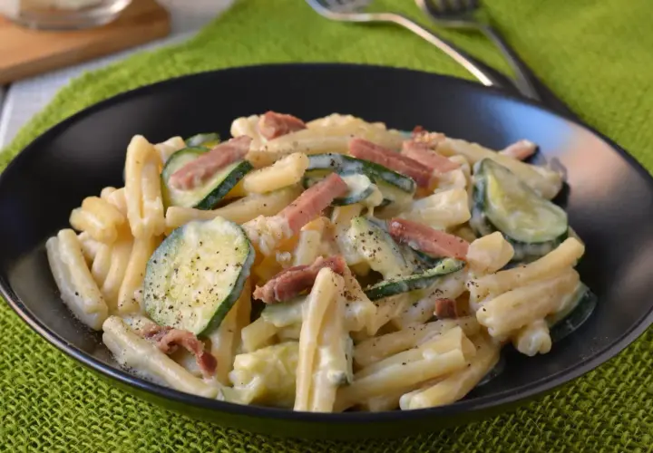 Piatto di pasta cremosa con zucchine a rondelle e cubetti di speck croccante, guarnito con pepe nero macinato.