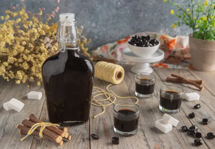 Bottiglia di liquore alla liquirizia scuro, bicchierini, bastoncini di liquirizia, zollette di zucchero e caramelle nere su un tavolo di legno.