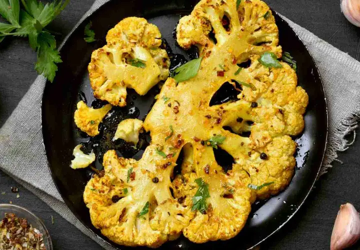 Fette di cavolo al forno dorate e speziate, servite su una padella nera con erbe fresche e pepe nero.