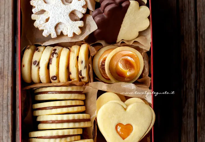 Scatola piena di biscotti classici: a fiocco di neve, con gocce di cioccolato, ripieni di caramello, a forma di cuore e ricoperti di cioccolato.