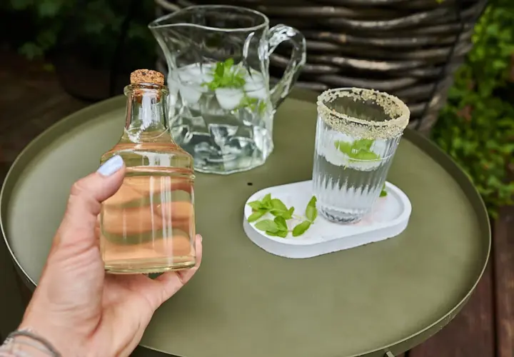 Mano tiene una bottiglietta di sciroppo di menta, accanto a un bicchiere e una caraffa con acqua, ghiaccio e foglie di menta.