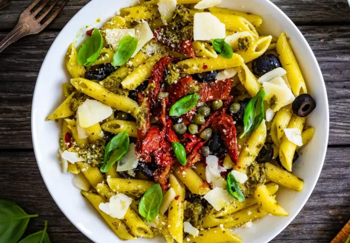 Penne al pesto con pomodori secchi, olive nere, capperi e scaglie di parmigiano. Un piatto colorato e saporito.