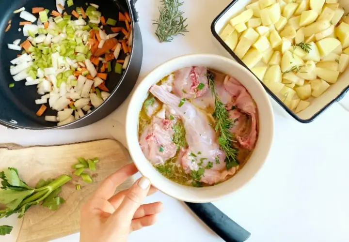 Preparazione per un coniglio alla cacciatora in bianco: pezzi di coniglio crudo in una ciotola, verdure tritate in padella e patate a cubetti in una teglia.