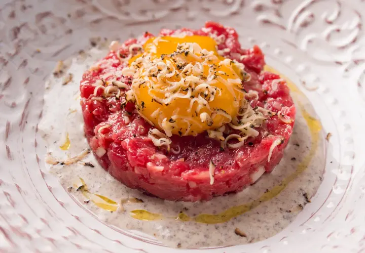 Deliziosa tartare di manzo finemente tritata, guarnita con tuorli d'uovo dorati e scaglie di formaggio, servita su un piatto elegante con una salsa cremosa.