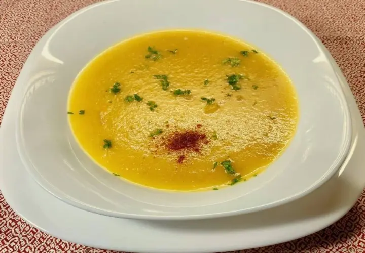 Una vellutata crema di peperoni, guarnita con prezzemolo fresco e una spolverata di paprika, servita in una ciotola bianca su un tovagliolo rosso.