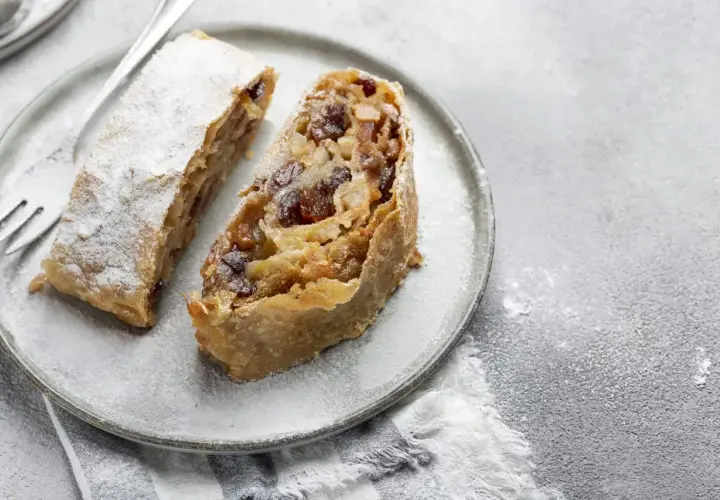 Due fette di strudel di mele con pasta sfoglia, cosparse di zucchero a velo, su un piatto grigio con una forchetta.
