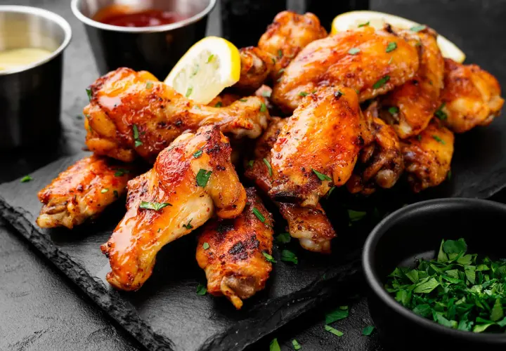 Alette di pollo dorate e succulente, guarnite con prezzemolo fresco e spicchi di limone, servite su una pietra scura con salse a lato.