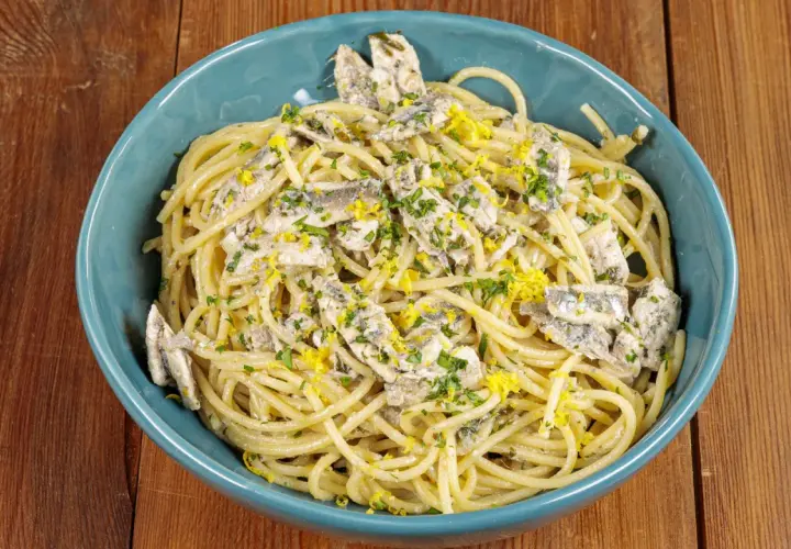 Spaghetti con acciughe, prezzemolo tritato e scorza di limone grattugiata in una ciotola blu su un tavolo di legno.