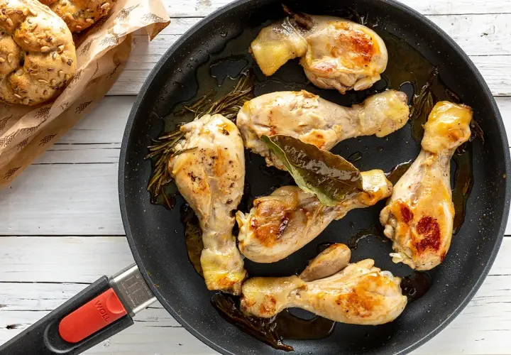 Fusi di pollo in padella, dorati e succulenti, con rosmarino e alloro. Accanto, panini integrali appena sfornati.