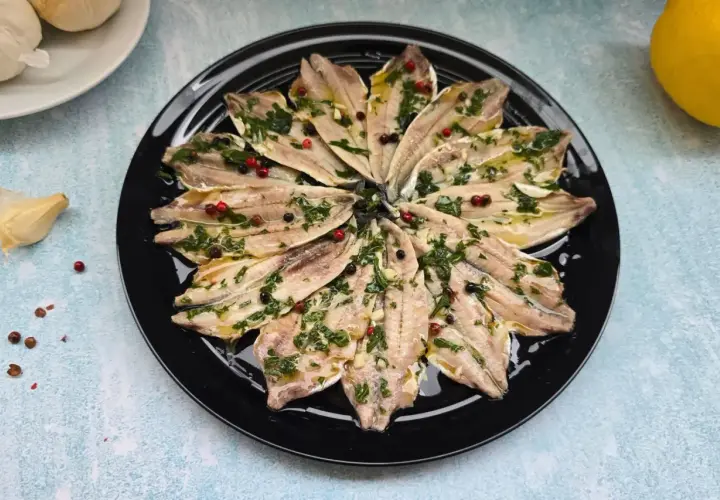 Alici marinate ricetta: filetti di acciughe conditi con erbe aromatiche, aglio e pepe in grani su un piatto nero.