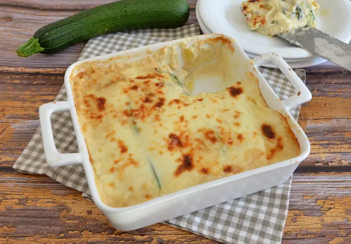 Parmigiana di zucchine bianca senza besciamella, dorata e filante, pronta per essere gustata. Accanto, una zucchina fresca.