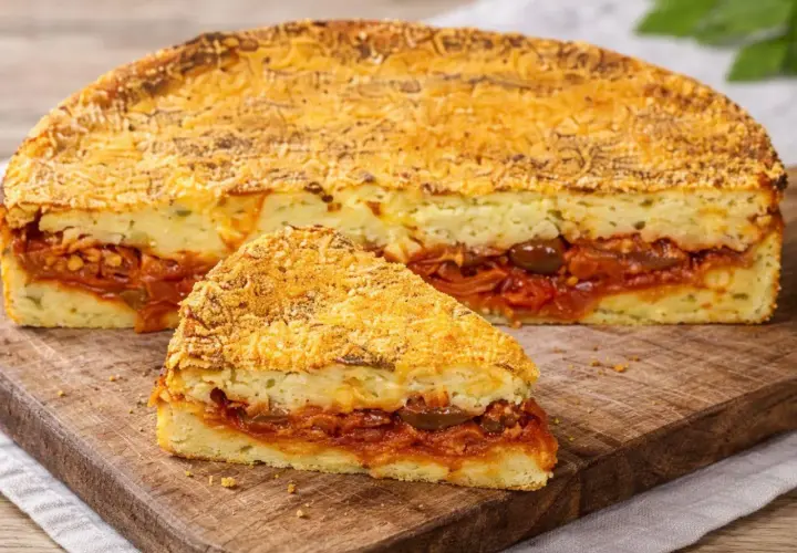 Una fetta di pitta di patate, con strati di purè e un ricco ripieno di pomodoro e olive, su un tagliere di legno.