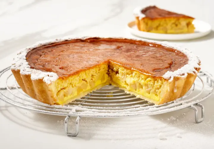 Torta di mele con crema pasticcera, con una fetta tagliata, cosparsa di zucchero a velo su una griglia metallica.
