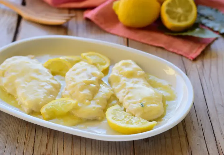 Scaloppine al limone cremose, succulente e profumate, guarnite con spicchi di limone fresco su un piatto bianco.