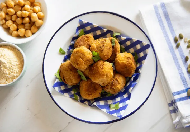 Piatto di polpette di ceci dorate e croccanti, guarnite con prezzemolo fresco. Accanto, ciotole con ceci e farina di ceci.