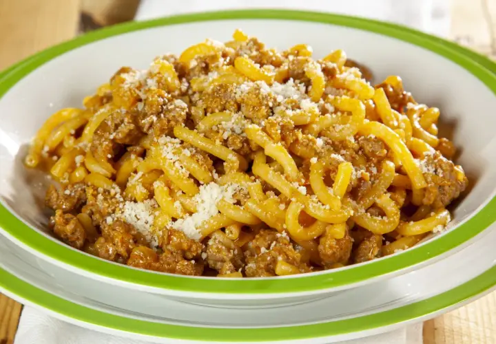 Piatto di gramigna alla salsiccia, condita con ragù e parmigiano grattugiato, servita in una ciotola bianca con bordo verde.